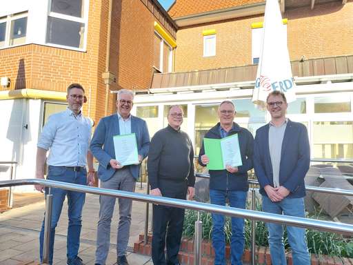 Fünf Männer stehen nebeneinander auf einer gepflasterten Fläche vor einem Gebäude aus roten Ziegelsteinen mit weißen Fensterrahmen. Zwei der Männer halten grüne Mappen in den Händen. Rechts im Bild befindet sich ein großer Sonnenschirm mit der teilweise sichtbaren Aufschrift „ERD“. Die Männer stehen hinter einem silbernen Geländer. Bild anklicken um zum kompletten Artikel zu gelangen.