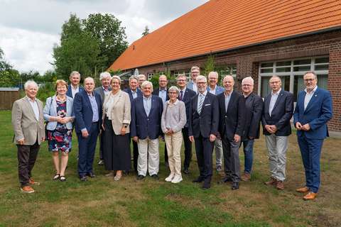 Gruppenfoto von etwa 20 Personen in zwei Reihen auf einer Wiese vor einem Backsteingebäude mit rotem Ziegeldach. Die meisten tragen elegante Kleidung wie Sakkos und Blazer. Alle blicken lächelnd in die Kamera.