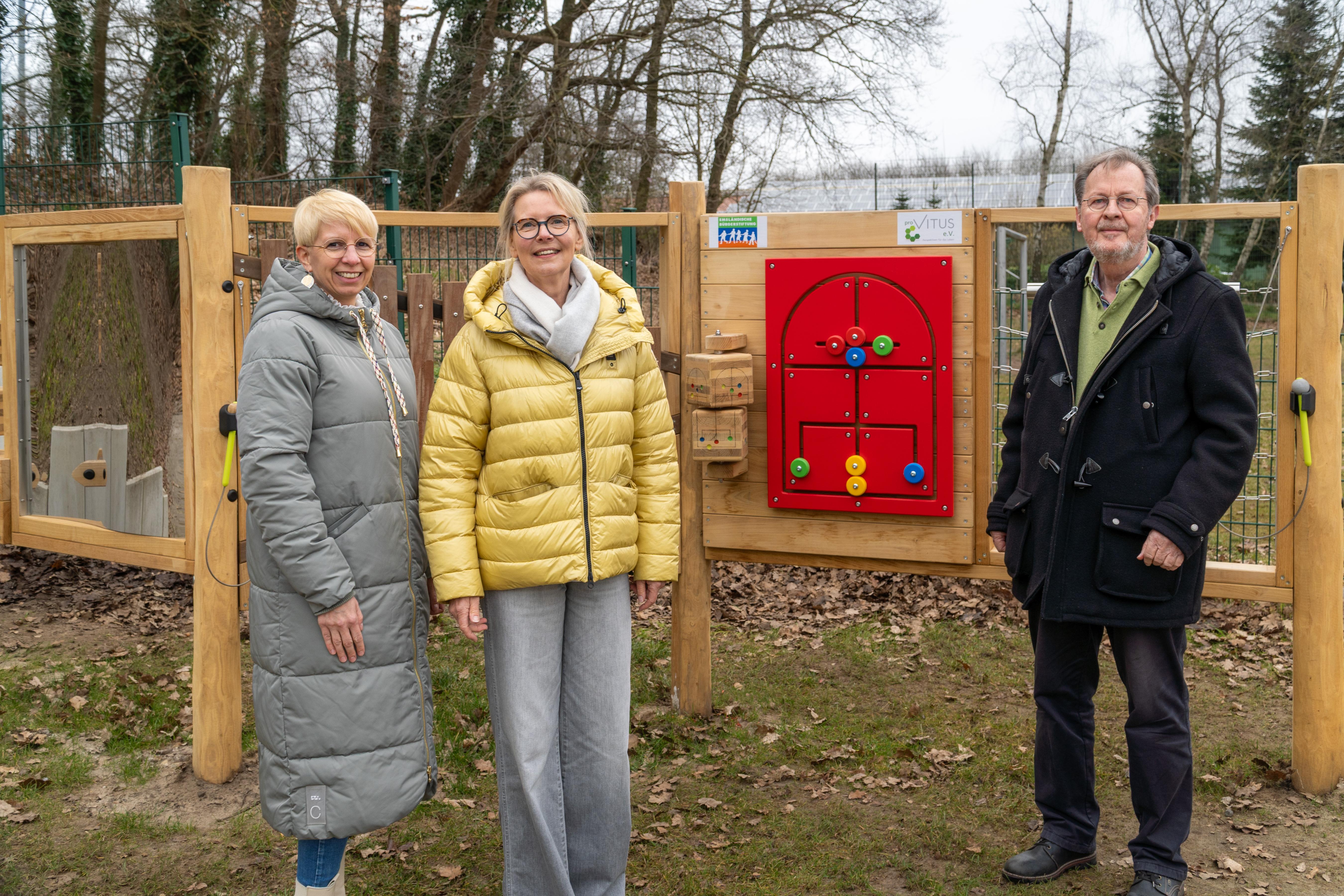 Drei Personen stehen im Freien vor einer bunt gestalteten Holzspielwand. Zwei Frauen tragen helle Winterjacken – eine in Grau, die andere in Gelb –, während der Mann rechts in dunkler Kleidung gekleidet ist. Die Wand hinter ihnen ist mit verschiedenen spielerischen Elementen ausgestattet, darunter ein rotes Element mit bunten Drehscheiben und Griffen. Auf der oberen rechten Seite befindet sich ein Schild mit dem Logo von „pro Vitus e.V.“. Im Hintergrund sind kahle Bäume und ein Maschendrahtzaun zu erkennen, was auf die kalte Jahreszeit hindeutet. Der Boden ist mit Laub bedeckt.