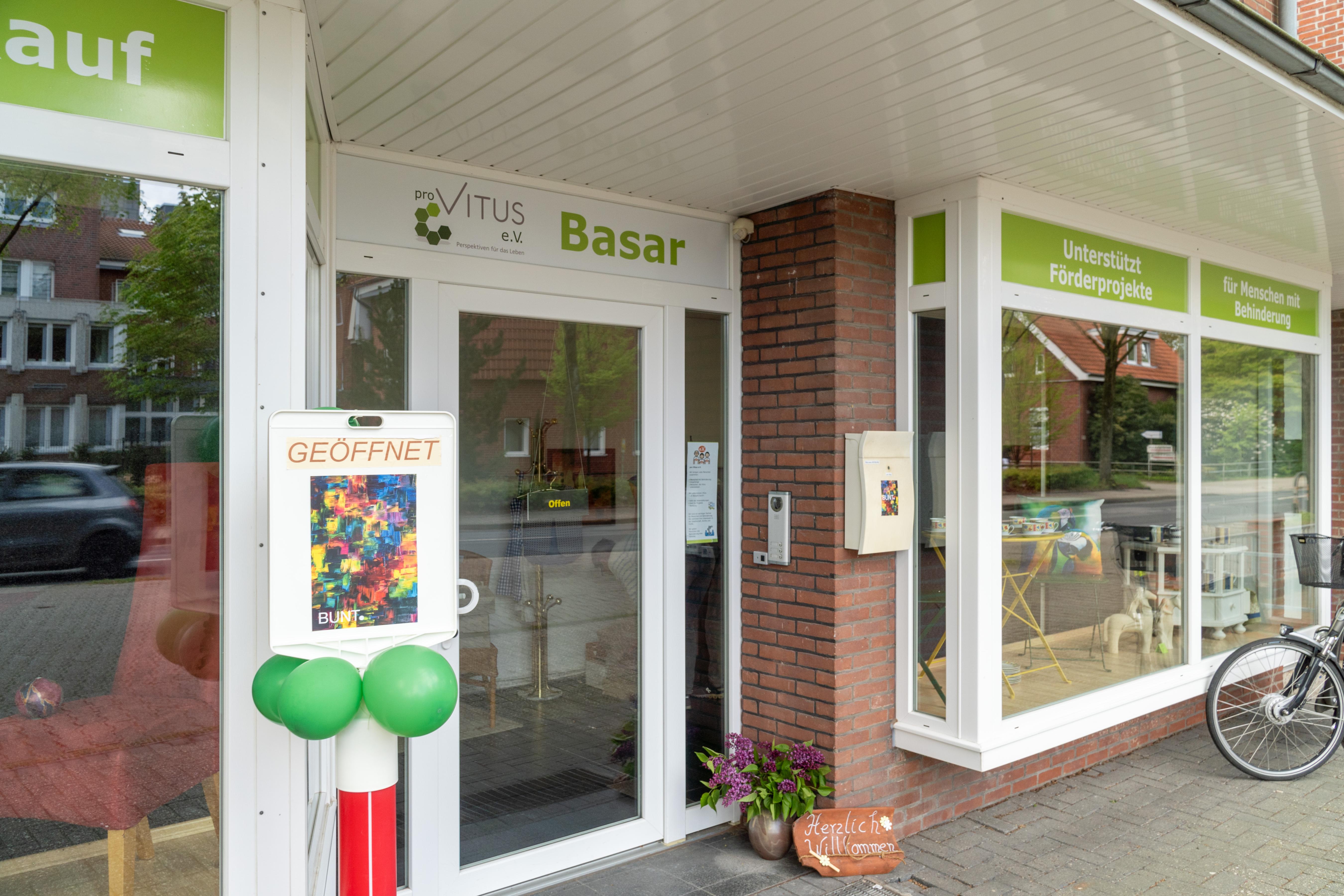 Das Bild zeigt den Eingang eines Ladens mit dem Namen „proVITUS e.V. Basar“. Über den Fenstern befinden sich grüne Schilder mit dem Hinweis, dass der Laden Projekte für Menschen mit Behinderungen unterstützt. Vor dem Eingang steht ein Schild mit der Aufschrift „GEÖFFNET“, darauf ist ein buntes abstraktes Gemälde zu sehen. An dem Schild sind drei grüne Luftballons befestigt. Der Eingang besteht aus Glasfenstern und Glastüren. Neben der Tür steht ein Blumentopf mit lila Blumen. Am rechten Bildrand ist teilweise ein Fahrrad zu erkennen.