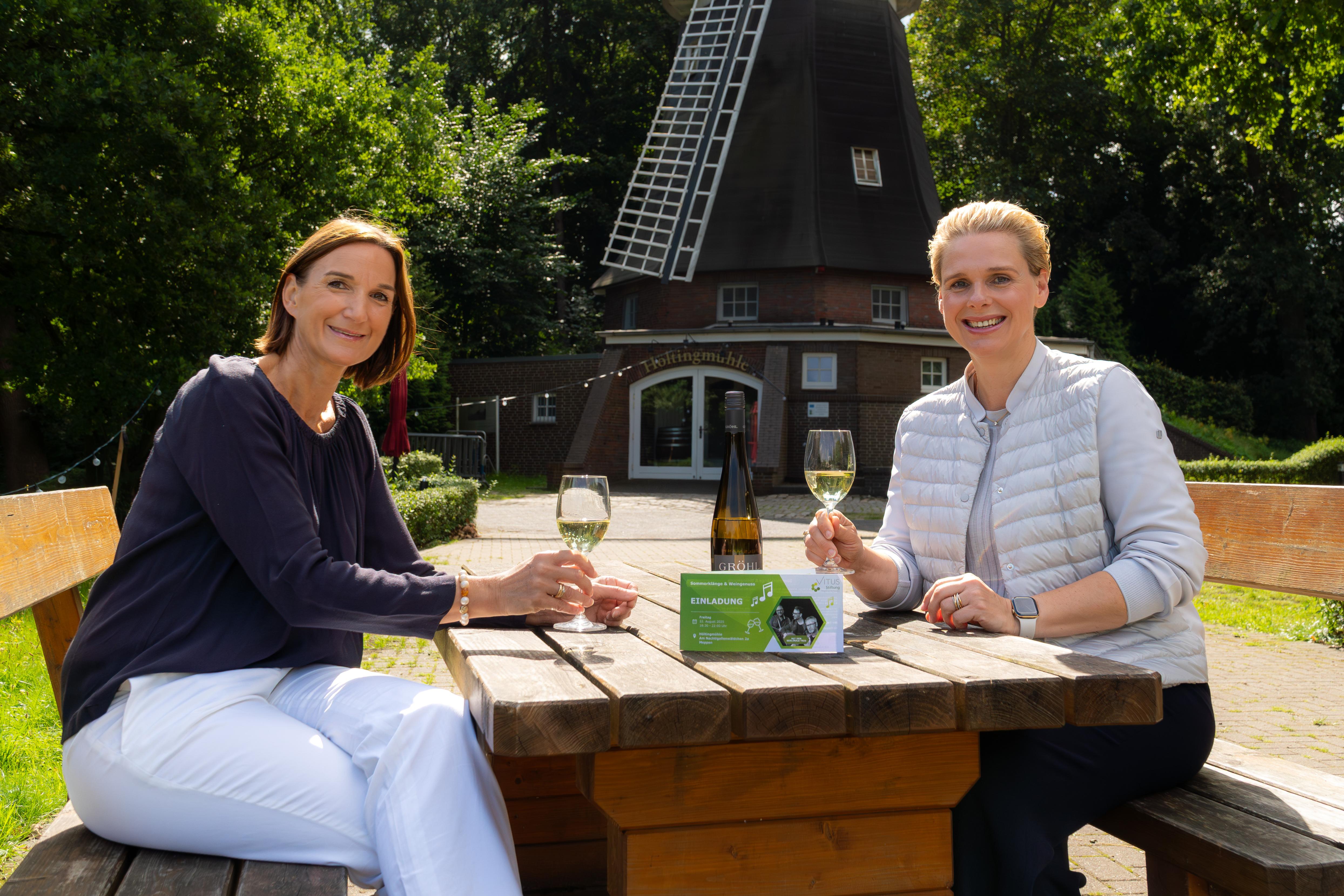 Das Bild zeigt zwei Personen, die an einem hölzernen Picknicktisch im Freien sitzen. Im Hintergrund ist eine Windmühle zu sehen, auf deren Gebäude der Schriftzug „Hofingmühle“ steht. Beide Personen halten ein Glas Weißwein in der Hand. Auf dem Tisch befinden sich eine Weinflasche und eine Einladungskarte, auf der unter anderem das Wort „EINLADUNG“ zu erkennen ist